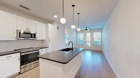Located at 2403 Mall Dr in North Charleston, SC, this thoughtfully designed one-bedroom, one-bathroom residence offers a modern and efficient layout across a single floor. The open-concept kitchen and living area serves as the heart of the home, anchored by a central island with a dark countertop, black matte faucet, and undermount sink. Globe pendant lights hang above the island, adding a stylish focal point. The kitchen is equipped with stainless steel appliances, white flat-front cabinetry, and a classic subway tile backsplash, delivering a clean, contemporary aesthetic. Light hardwood-style flooring flows throughout the main living areas, complementing the bright, neutral color palette. The living area benefits from natural light through large windows and a glass-panel door that opens to a private outdoor space, accompanied by a ceiling fan overhead. The bedroom is carpeted for comfort and features a ceiling fan, a window with natural views, and direct access to the bathroom. The bathroom includes a white vanity with dark countertop, black hardware accents, and hardwood-style flooring. A walk-in closet with multi-tier wire shelving provides ample organized storage, completing this well-appointed residence.