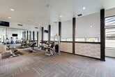 Located at 9600 Golf Lakes Trail in Dallas, TX, this fitness center offers a generously sized, single-floor workout facility designed to accommodate a full range of exercise needs. The space features a bright, open layout enhanced by large windows along the exterior walls, inviting abundant natural light and providing pleasant views of the surrounding landscape, including a nearby body of water. The vaulted ceiling in one section adds a sense of vertical spaciousness, while recessed lighting throughout ensures a well-lit environment at all hours.

The interior is finished with multi-tone patterned carpet tiles that lend a contemporary aesthetic to the floor. Bold dark structural columns punctuate the space, complemented by a textured herringbone accent wall panel that adds visual interest. Full-length mirrors line one wall, enhancing the sense of depth and allowing exercisers to monitor their form.

The facility is outfitted with a comprehensive selection of equipment, including cable and resistance machines, adjustable benches, a dumbbell rack, treadmills, recumbent bikes, and stair climbers. A dedicated area features medicine balls and stability balls on a organized storage rack for functional fitness training. A water fountain is conveniently available within the space, and access points connect to adjacent amenity areas, making this a complete and thoughtfully designed fitness destination.