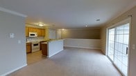 This single-floor space features an open-concept layout that seamlessly connects the kitchen and living area, creating a cohesive and functional environment. The kitchen is outfitted with warm honey-toned wood cabinetry, a white electric range, and a built-in microwave above the stove. Light tile flooring runs throughout the kitchen area, providing a clean and practical surface. A peninsula counter serves as a natural divider between the kitchen and the adjacent living space, offering both additional workspace and a visual transition between the two areas. The living area is finished with plush wall-to-wall carpeting in a neutral tone, lending warmth and comfort underfoot. Crown molding runs along the ceiling perimeter throughout the space, adding a refined architectural detail. The walls are painted in a soft, warm taupe that complements the overall neutral palette. Recessed lighting and flush-mount ceiling fixtures provide consistent illumination throughout. A doorway visible from the kitchen suggests additional rooms or utility spaces beyond. The overall ambiance is clean, understated, and move-in ready, suited for everyday living.