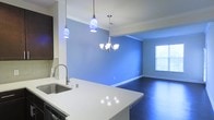 This single-floor space offers an open-concept layout that seamlessly connects the kitchen and dining areas, creating a cohesive and inviting living environment. The kitchen features a sleek white countertop island with a stainless steel sink, dark cabinetry with a tile backsplash, and a set of contemporary blue pendant lights that add a vibrant pop of color overhead. The transition into the dining area is marked by a sophisticated chandelier with classic white shades, providing warm, ambient lighting. Bold blue accent walls give the space a distinctive and energetic character, complemented by crisp white trim and baseboards throughout. Dark hardwood flooring runs across the dining area, adding depth and contrast to the bright walls and countertops. A window along the far wall allows natural light to filter into the space, enhancing its open and airy feel. The overall design balances modern fixtures with bold color choices, resulting in a space that is both functional and visually dynamic.