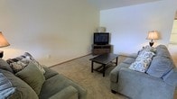 This single-story space features a living area that serves as the central gathering space, designed with simplicity and comfort in mind. The room is finished with wall-to-wall carpeting in a neutral beige tone, which adds warmth underfoot and contributes to the overall relaxed atmosphere. The walls are painted in a soft, light neutral color, creating a clean and airy backdrop that allows for flexible furnishing and décor choices.

The living area is furnished with two grey upholstered sofas arranged to face a corner media console, which houses a flat-screen television and offers drawer storage for media accessories. A dark-finished rectangular coffee table anchors the seating arrangement, providing a functional centerpiece to the space. The corner placement of the entertainment setup makes efficient use of the room's layout.

The ceiling height adds a sense of openness to the room, with the upper wall space left unadorned, contributing to a spacious feel. Baseboard trim runs along the perimeter of the walls, adding a subtle architectural detail. Overall, the space presents a straightforward, functional layout well-suited for comfortable daily use.