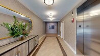 Located at 443 Duane St in Glen Ellyn, Illinois, this single-floor lobby corridor presents a carefully curated, upscale aesthetic that sets a welcoming tone upon entry. The space is defined by its light-toned herringbone-patterned flooring, accented by darker wood-toned border inlays that draw the eye down the length of the corridor, creating a sense of depth and intentional design.

The walls are finished in a subtly textured, neutral-toned wallcovering adorned with a delicate pattern, complemented by crisp white crown molding that adds architectural refinement. Flush-mount light fixtures with warm, diffused illumination line the ceiling, maintaining an inviting ambiance throughout.

A standout decorative vignette anchors one side of the corridor, featuring a stylish credenza topped with a lush floral arrangement and a gold-framed ornate mirror that reflects the space elegantly. Curated artwork punctuates the far end of the hallway, adding a gallery-like quality.

The entry vestibule includes floor-to-ceiling glass doors and sidelights that allow natural light to filter in, offering views to the exterior. A stainless steel elevator with polished doors provides vertical access, seamlessly integrated into the corridor's refined design. Overall, the space exudes a polished, boutique-style character.