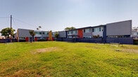 Located in San Diego, CA, this outdoor dog park offers a generously sized, well-kept grassy lawn enclosed by sturdy black chain-link fencing, providing a safe and open environment for pets and their owners. The park is framed by a vibrant residential community featuring modern two-story buildings with bold color accents — including deep navy blue, bright red, and warm orange — contrasting against clean gray siding and large windows that reflect the sunny Southern California climate. A covered pergola sits adjacent to the lawn, offering a shaded gathering spot for visitors. Benches are positioned throughout the perimeter, providing comfortable seating while pets enjoy the open green space. BBQ stations are also available near the park's edge, adding a recreational amenity for residents. The park benefits from abundant natural light under clear blue skies, typical of the San Diego area. Mature trees dot the surroundings, offering additional shade and greenery. The space is accessed through a gated entry, ensuring security for both pets and visitors. Surrounding views include open skies and palm trees characteristic of the coastal San Diego landscape, making this dog park a functional and inviting community amenity.