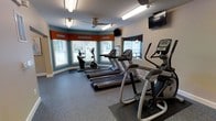 This fitness center offers a functional and inviting workout environment highlighted by teal accent walls and warm orange valances that add a distinctive decorative touch. The space is illuminated by a combination of fluorescent ceiling fixtures and a ceiling fan, ensuring consistent lighting and airflow throughout workouts. Multiple windows fitted with white blinds line the walls, allowing natural light to filter in while maintaining privacy. The durable rubber flooring provides excellent traction and cushioning underfoot, ideal for high-impact exercise. Cardio equipment is the focal point of the room, featuring a row of treadmills, an upright stationary bike, and an elliptical cross-trainer, all positioned to face the windows for an engaging workout experience. Wall-mounted televisions are strategically placed for easy viewing from the equipment, adding entertainment options during exercise sessions. The layout is thoughtfully organized to maximize the available floor space while maintaining comfortable clearance between machines. Informational signage is posted on the walls, providing guidance on equipment use and facility rules. Overall, the space maintains a clean, functional aesthetic suited to daily fitness activities.