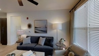 The living area of this single-floor space is designed with a balance of comfort and refined aesthetics. The room centers around a substantial charcoal-blue sofa adorned with an assortment of textured throw pillows in neutral tones, providing a welcoming focal point. A ceiling fan overhead contributes both function and a subtle architectural detail to the space.

The walls are finished in a clean, light neutral tone that enhances the room's brightness. A framed black-and-white photographic print of a suspension bridge, set in a warm gold frame, serves as a standout decorative element, complemented by a sunburst mirror mounted nearby. Together, these pieces add visual interest and a curated sensibility to the room.

Lighting is layered thoughtfully throughout the space. A tall floor lamp with a large white drum shade provides warm ambient light, while additional accent lighting contributes to the room's intimate feel. A large window dressed with horizontal blinds and neutral linen drapes on a black rod allows natural light to filter in, brightening the space while maintaining privacy. A small side table with a floral arrangement and personal framed photos adds a personalized, lived-in charm to the overall setting.