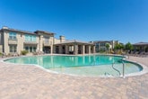 Located at 5300 Antequera Rd NW in Albuquerque, New Mexico, this outdoor amenity space offers a resort-style experience designed for relaxation and socializing under the expansive New Mexico sky. The centerpiece is a generously sized swimming pool with a smooth paver surround, lined with lounge chairs that invite residents to soak up the abundant Southwest sunshine. The pool area is framed by mature trees and lush landscaping, providing a pleasant contrast to the warm, stucco-and-stone architecture of the surrounding multi-story buildings. Adjacent to the pool is a covered outdoor pavilion that serves as an elegant gathering space. Stone-clad columns support the overhead structure, which is equipped with ceiling fans to ensure comfortable airflow during warm days. The pavilion is furnished with plush, well-appointed seating arrangements set atop decorative area rugs, creating a welcoming lounge atmosphere. A wall-mounted flat-screen television and a stone-surround fireplace make this covered area equally functional for cooler evenings. A bar-height seating counter adds a social dining element to the space. Access to the pool deck is provided through a secured wrought iron gate, ensuring privacy and safety. The cohesive paver flooring throughout ties the entire outdoor space together with a refined, upscale aesthetic.