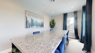This single-floor meeting space is thoughtfully designed to support professional gatherings in a clean, modern setting. The centerpiece of the room is a generously sized conference table finished in a light granite surface with subtle grey and white veining, providing both an elegant and functional workspace. Surrounding the table are coordinated blue upholstered chairs that add a pop of color against the neutral, light grey walls.

Recessed ceiling lighting supplements the natural light that filters through multiple windows dressed with teal-toned grommet-style curtains, which can be drawn to control light and privacy as needed. The carpeted floor adds a layer of warmth and acoustic comfort to the space.

A large framed abstract artwork in blue and grey tones anchors one wall, complementing the room's overall color palette and contributing to a polished, curated ambiance. A tall fiddle-leaf fig plant adds a natural, organic element to the corner of the room. The walls are finished with clean white baseboards, and built-in electrical outlets along the baseboard offer convenient connectivity for presentations or devices. Overall, the space strikes a balance between functionality and refined aesthetic, making it well-suited for meetings, presentations, or collaborative work sessions.
