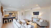 This well-appointed one-bedroom, one-bathroom residence showcases a clean, contemporary design anchored by light wood flooring and a crisp white palette throughout. The open-concept living area is filled with natural light from a large picture window framing urban views, and features recessed lighting and a tray ceiling detail that adds architectural depth. The living space flows seamlessly into the kitchen, which is outfitted with two-tone cabinetry in light wood and grey, stainless steel appliances including a French door refrigerator and gas range, a built-in microwave, and a peninsula with a quartz countertop and bar seating — ideal for casual dining or entertaining. The bedroom is a serene retreat with its own large window offering city views, a wall-mounted television, access to a dedicated closet, and tray ceiling detailing that mirrors the living area. The bathroom is finished in neutral tones with tile flooring, a floating white vanity with a brushed gold-framed mirror, and a full tub-shower combination. A separate closet provides additional storage. Throughout the residence, the cohesive modern aesthetic, abundant natural light, and urban outlook create a comfortable and stylish living environment.