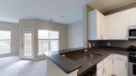 Located at 900 Reisling Lane in West Chester, PA, this single-floor residence offers a thoughtfully designed open-concept layout that seamlessly connects the kitchen and living areas. The kitchen is outfitted with crisp white cabinetry, sleek dark countertops, and a stainless steel undermount sink, creating a striking contrast that gives the space a modern, polished aesthetic. A peninsula-style counter extends into the living area, providing additional workspace and a natural gathering point. The living area is bright and airy, benefiting from a large multi-pane window and a glass door that bathe the space in natural light while offering views of the outdoor surroundings. Light-colored carpeting throughout the living area adds warmth and comfort underfoot. Neutral wall tones and clean lines throughout contribute to a calm, welcoming ambiance. The single-floor layout ensures easy accessibility and a cohesive flow between spaces, making everyday living both practical and comfortable. This residence in West Chester, PA combines functional design with contemporary finishes for a refined and inviting living experience.