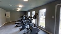 This fitness center offers a dedicated space for cardiovascular training and exercise. The room is outfitted with a lineup of modern cardio machines, including ellipticals and treadmills from Life Fitness, arranged in a row to maximize floor space and accessibility. Large double-hung windows line the exterior wall, flooding the room with natural light and providing views of the surrounding outdoor greenery, creating a bright and motivating workout environment. The neutral gray walls and light-toned flooring contribute to a clean, contemporary aesthetic that keeps the focus on functionality. Overhead, flush-mount ceiling lights provide consistent, even illumination throughout the space, ensuring visibility during any time of day. The textured ceiling adds a subtle architectural detail that complements the overall design. The single-floor layout ensures ease of access, making the space practical and straightforward to navigate. Overall, this fitness center strikes a balance between a well-maintained, professional atmosphere and an inviting, comfortable setting suitable for a range of fitness routines.
