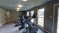 This fitness center offers a dedicated space for cardiovascular training and exercise. The room is outfitted with a lineup of modern cardio machines, including ellipticals and treadmills from Life Fitness, arranged in a row to maximize floor space and accessibility. Large double-hung windows line the exterior wall, flooding the room with natural light and providing views of the surrounding outdoor greenery, creating a bright and motivating workout environment. The neutral gray walls and light-toned flooring contribute to a clean, contemporary aesthetic that keeps the focus on functionality. Overhead, flush-mount ceiling lights provide consistent, even illumination throughout the space, ensuring visibility during any time of day. The textured ceiling adds a subtle architectural detail that complements the overall design. The single-floor layout ensures ease of access, making the space practical and straightforward to navigate. Overall, this fitness center strikes a balance between a well-maintained, professional atmosphere and an inviting, comfortable setting suitable for a range of fitness routines.
