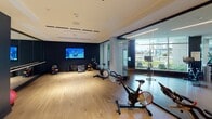 Located on Element Way in Charlotte, NC, this single-floor fitness center offers a well-appointed, contemporary exercise facility designed to support a wide range of workout routines. The space is divided into distinct functional zones, each thoughtfully equipped and finished to a high standard.

The cycling area features multiple stationary bikes arranged on smooth light wood flooring, complemented by bold teal-painted accent walls and floor-to-ceiling glass partitions that visually open the space. Large windows along one side invite natural light and provide outdoor views, enhancing the workout environment. A wall-mounted flat-screen TV is positioned for easy viewing during cardio sessions.

A dedicated functional training zone showcases wall-mounted resistance systems, including TRX-style suspension trainers and resistance bands anchored to a structured wall rig. Mirrored walls expand the sense of space and allow for form monitoring during exercises.

A strength and free weights area is stocked with a full rack of dumbbells, exercise mats, medicine balls, and a stability ball. Additional cardio equipment, including stationary bikes and rowing machines, rounds out the offerings. Recessed and track lighting throughout provides consistent, well-distributed illumination, creating a clean, professional atmosphere suitable for focused training.