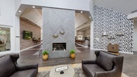 This thoughtfully designed single-floor community space makes a striking first impression with its soaring vaulted ceilings and an abundance of natural light streaming through floor-to-ceiling windows and a grand arched window at the entry. The centerpiece of the main lounge is a dramatic floor-to-ceiling fireplace clad in chevron-patterned grey tile, flanked by lush greenery and framed by rich wood-look flooring laid in a herringbone pattern. The open-concept layout flows seamlessly between multiple seating areas, including a generously sized sectional leather sofa arrangement anchored by a patterned area rug and a wall-mounted flat-screen television. An eye-catching black-and-white abstract accent wall adds a bold, graphic element to the interior design. A curved sunroom-style lounge with a coffered ceiling and panoramic windows offers a bright, inviting retreat with views of the surrounding greenery. The kitchen area is well-appointed with grey cabinetry, a herringbone-tiled backsplash, dark countertops, stainless steel appliances, and a bar-height island illuminated by a row of industrial-style pendant lights. Additional corridors and transition spaces are clean and welcoming, featuring tiled flooring, warm accent seating, and modern cable railings along the stairwell. Throughout, the space balances contemporary design with a warm, approachable atmosphere.