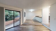 This single-floor residence has been thoughtfully updated throughout, showcasing a clean, modern aesthetic with wood-look vinyl plank flooring running consistently across the living areas. The spacious living room serves as a central gathering space, anchored by a tiled fireplace surround and flanked by a large window and a sliding glass door that opens to a private outdoor patio, flooding the room with natural light and views of surrounding greenery.

Adjacent to the living room, a raised dining or flex area provides a subtle division of space while maintaining an open connection to the kitchen. The kitchen is well-equipped with white shaker cabinetry, dark countertops, stainless steel appliances including a refrigerator, electric range, and dishwasher, and a deep undermount sink with a modern faucet.

The bedroom is generously sized with a window providing natural light and ample wall space. Two bathrooms serve the unit: a full bath featuring a tub-shower combination, a vanity with white cabinetry, and a separate water closet. Textured ceilings and crisp white walls complete the clean, updated interior throughout.
