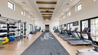 Located in San Tan Valley, AZ, this fitness center offers an impressively spacious and thoughtfully designed exercise facility. The single-floor layout is anchored by soaring ceilings adorned with rustic exposed wood beams and ceiling fans, creating a bright, open, and inviting atmosphere. Rows of large black-framed windows line the walls on two levels, flooding the interior with natural light and providing views of the surrounding community and landscaping.

Light wood-style flooring runs throughout the space, complemented by a central branded turf runner that divides the floor into distinct workout zones. The cardio section is well-equipped with multiple treadmills, stair climbers, rowing machines, and stationary cycling bikes, each outfitted with personal screens. Wall-mounted televisions provide additional entertainment throughout.

The strength training area features a robust multi-functional cable and power rack system, a full dumbbell rack, weight plates, kettlebells, medicine balls, and benches, accommodating a wide range of resistance training exercises. Large mirrors are strategically placed to maximize perceived space and assist with form. Direct exterior access through glass-paned doors connects the gym to an outdoor area. The overall ambiance is modern and clean, combining a contemporary aesthetic with a welcoming community fitness environment.