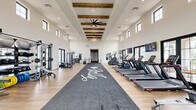 Located in San Tan Valley, AZ, this fitness center offers an impressively spacious and thoughtfully designed exercise facility. The single-floor layout is anchored by soaring ceilings adorned with rustic exposed wood beams and ceiling fans, creating a bright, open, and inviting atmosphere. Rows of large black-framed windows line the walls on two levels, flooding the interior with natural light and providing views of the surrounding community and landscaping.

Light wood-style flooring runs throughout the space, complemented by a central branded turf runner that divides the floor into distinct workout zones. The cardio section is well-equipped with multiple treadmills, stair climbers, rowing machines, and stationary cycling bikes, each outfitted with personal screens. Wall-mounted televisions provide additional entertainment throughout.

The strength training area features a robust multi-functional cable and power rack system, a full dumbbell rack, weight plates, kettlebells, medicine balls, and benches, accommodating a wide range of resistance training exercises. Large mirrors are strategically placed to maximize perceived space and assist with form. Direct exterior access through glass-paned doors connects the gym to an outdoor area. The overall ambiance is modern and clean, combining a contemporary aesthetic with a welcoming community fitness environment.
