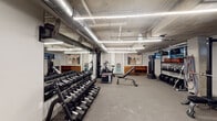 This fitness facility presents a well-appointed, industrial-style gym space spread across a single level. The exposed concrete ceiling with large-diameter silver ductwork and linear LED lighting creates a raw, functional aesthetic throughout. The flooring consists of durable, neutral-toned carpet-style gym material suited for high-traffic use.

The main workout area is generously sized and organized into distinct zones. A full dumbbell rack lines one wall, accompanied by a large floor-to-ceiling mirror that visually expands the space and aids in form monitoring. Warm-toned wooden storage cubbies add a subtle contrast to the otherwise industrial palette. Cardio equipment includes stationary bikes, a treadmill, and a rowing machine, while strength training is further supported by a cable machine, adjustable bench, kettlebell rack, weighted plates, a punching bag, and a plyo box.

A secondary, glass-partitioned room contains additional equipment, including a treadmill and rowing machine, offering a more private training environment. Abstract artwork adorns select walls, softening the utilitarian character of the space. A built-in water station provides added convenience. Industrial pedestal fans are positioned throughout to support ventilation and airflow, complementing the exposed mechanical systems overhead.