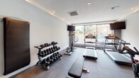This private fitness space is thoughtfully designed to deliver a complete workout experience within a sleek, modern aesthetic. The room features large-format tile flooring in a neutral gray tone, recessed lighting set into a smooth white ceiling, and crisp white walls that contribute to a bright, clean atmosphere.

A floor-to-ceiling glass window wall along one side floods the space with natural light while offering views of the surrounding outdoor landscape, creating an open, energizing environment. A full-width wall mirror on the opposite side visually expands the room and allows for form monitoring during exercise.

The space is well-equipped with a comprehensive range of fitness equipment, including treadmills, upright and spin cycles, a multi-station cable machine, and a tiered dumbbell rack holding a full set of free weights. Stability balls are neatly stored on a wall-mounted rack, and a punch bag is mounted nearby for conditioning work. Wall-mounted decorative weights add a stylish visual accent.

A glass partition wall connects the fitness area to an adjacent living space, maintaining a sense of openness while defining each zone. The overall ambiance is professional and polished, combining functional gym design with contemporary interior finishes.