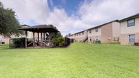 This property features a spacious outdoor common area that serves as a central gathering space for residents. The grounds are well-kept, with a broad, lush green lawn that provides ample open space for leisure and recreation. A prominent covered gazebo with a dark wood frame and a pitched roof anchors the outdoor area, offering a shaded retreat enclosed by wrought iron fencing and surrounded by decorative landscaping. The gazebo provides a comfortable space for outdoor socializing and relaxation. Flanking the green space are multi-unit residential buildings constructed of brick in a neutral tan tone, with exterior staircases providing access to upper levels. Each building features evenly spaced windows and individual HVAC units, reflecting a functional and practical design. The overall layout of the grounds emphasizes open space and accessibility, with the buildings set back from the central lawn to create a sense of openness. Mature trees visible in the background contribute to a natural, established feel, while the well-maintained landscaping throughout reflects consistent upkeep of the property's exterior.