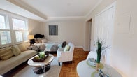 This single-floor studio is designed to maximize its open-concept layout, combining the sleeping and living areas into one cohesive, functional space. The warm parquet hardwood flooring adds a timeless character to the room, complementing the neutral beige walls and crisp white crown moulding that frames the ceiling, lending a polished, classic aesthetic throughout.

The sleeping area is anchored by a generously sized bed dressed in decorative bedding, positioned beside a bedside lamp that adds a warm, ambient glow. Adjacent to it, a seating arrangement featuring white upholstered armchairs and a round coffee table creates a distinct lounge area, ideal for relaxing or entertaining. A soft area rug underneath defines this zone and adds texture to the space.

Near the foreground, a small dining or accent table is styled with decorative greenery, bringing a touch of nature indoors. A closet with bifold doors is visible near the entryway, offering practical storage solutions. The overall ambiance is calm and inviting, characterized by its well-coordinated furnishings, clean lines, and thoughtful use of space.