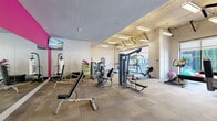 Located at 116 Holmes Ave NW in Huntsville, Alabama, this fitness center offers a comprehensive workout environment designed to accommodate a wide range of exercise routines. The space is thoughtfully laid out across a single floor, with distinct zones dedicated to strength training, cardio, and functional fitness.

The strength training area is well-stocked with multiple cable machines, weight benches, and resistance equipment, providing options for both isolated and compound movements. A bold magenta accent wall adds a vibrant visual focal point, complemented by a wall-mounted television for entertainment during workouts. Full-length mirrors along one wall enhance the sense of space while allowing users to monitor their form.

The cardio section includes elliptical trainers, a rowing machine, a Peloton spin bike, and treadmills, positioned near large floor-to-ceiling windows that allow ample natural light and views to the outdoors. A medicine ball rack and additional functional training accessories round out the available equipment.

The industrial-modern aesthetic is defined by exposed concrete columns, suspended linear lighting fixtures, and a neutral-toned carpet tile floor throughout. Large glass partitions connect the fitness center visually to adjacent common areas, contributing to an open, airy atmosphere. Posted fitness center rules ensure an organized and safe environment for all users.