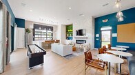 Located at 151 S Buddy Hayes Blvd in Anna, Texas, this single-story community clubhouse offers a well-appointed gathering space designed with modern aesthetics and functionality in mind. The open-plan layout is anchored by a vibrant teal accent wall that adds bold color contrast against crisp white finishes and light wood-look flooring throughout.

The main lounge area features a sleek electric fireplace set into a textured stone surround, flanked by a built-in display niche with a living moss wall and neon signage. A wall-mounted TV and comfortable seating arrangements create an inviting atmosphere for both casual relaxation and social events. A billiards table and shuffleboard table provide recreational options, while a banquette seating area with bistro tables and curated artwork along a textured wallpapered wall offers a more intimate setting.

The kitchenette is equipped with white shaker cabinetry, a stainless steel refrigerator, white countertops, and pendant lighting over a serving counter. A dramatic black-framed garage-style window floods the area with natural light while offering views to the outdoor patio. Sliding glass doors along the teal-accented wall open directly to an outdoor patio and pool area, seamlessly connecting the interior to the exterior amenities. The space also includes a closet for storage.