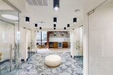 Located at 3100 Lake Villa Dr in Metairie, Louisiana, this thoughtfully designed professional space offers a refined blend of modern aesthetics and functional versatility. The single-floor layout encompasses a central open corridor anchored by a bold, patterned carpet that flows through the heart of the space, contrasting beautifully with the herringbone-tiled flooring found in the surrounding work and lounge areas.

Multiple glass-enclosed private rooms line the space, providing acoustically separate areas ideal for focused work or small meetings. Each glass office features sleek chrome hardware and is furnished with stylish seating in warm gold and muted jewel tones. Cylindrical black pendant lights are suspended from white ceilings throughout, delivering consistent, modern illumination across every zone.

A built-in kitchenette serves as a central amenity, showcasing warm wood cabinetry, open display shelving adorned with greenery, a sink, and a beverage refrigerator — all framed by large windows that invite natural light and exterior views. Adjacent seating areas feature marble-topped tables paired with plush purple and gold chairs, offering comfortable spaces for casual meetings or collaborative work.

Warm wood-paneled accent walls add texture and sophistication to the seating nooks, while a round upholstered ottoman in the central corridor provides a welcoming focal point. The overall ambiance is contemporary, polished, and professionally inviting.
