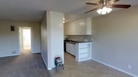 This single-story space features an open-concept layout that connects the main living area to the kitchen, creating a cohesive and functional floor plan. The kitchen is equipped with white upper cabinetry providing ample storage, dark countertops, a sink, and a dishwasher. A built-in white shelving unit serves as a subtle divider between the kitchen and the adjoining living space, adding both visual interest and practical storage. The living area is fitted with a ceiling fan with an integrated light fixture, ensuring comfortable air circulation and ambient lighting. Flooring transitions from tile in the kitchen and main area to carpet in an adjacent hallway or room, adding textural variety throughout. The walls are painted in a neutral gray tone, complementing the white trim and cabinetry. A textured popcorn ceiling is consistent throughout the space. A hallway visible in the background suggests additional rooms beyond the main area. Overall, the space presents a clean, straightforward aesthetic with practical features suited to everyday living.