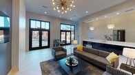 Located in the heart of downtown Hickory, North Carolina, this single-floor community lounge offers a sleek, contemporary design with thoughtful details throughout. The main gathering area features light-toned herringbone-patterned tile flooring and soaring ceilings that amplify the sense of openness. A striking sputnik-style chandelier serves as a dramatic focal point overhead, complementing the recessed lighting that illuminates the space evenly.

The lounge is furnished with a comfortable sectional sofa, accent chairs, and a low-profile coffee table arranged atop a plush area rug, creating an inviting social setting. A wall-mounted flat-screen television and additional seating nooks provide flexibility for both relaxed gatherings and focused use. Large black-framed windows and glass-paneled doors flood the interior with abundant natural light while offering views of and direct access to an outdoor patio area with bistro-style seating.

A built-in kitchenette lines one wall, featuring dark cabinetry with a wood-grain finish, a crisp white countertop with an undermount sink, and pendant lighting overhead. The neutral gray and white color palette, accented with warm yellow throw pillows and carefully selected décor, lends the space a polished yet welcoming ambiance throughout.