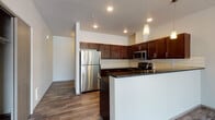 Located at 19112 East Alki Avenue in Spokane Valley, Washington, this single-floor unit offers a thoughtfully designed layout with modern finishes throughout. The open-concept living and kitchen area serves as the heart of the space, where wood-look flooring extends seamlessly underfoot. The kitchen is equipped with rich, dark-toned cabinetry, stainless steel appliances including a refrigerator, range, and microwave, and dark granite countertops complemented by a classic subway tile backsplash. Pendant lighting and recessed fixtures illuminate the space effectively. A breakfast bar adds functional counter seating and further connects the kitchen to the open living area.

The living area is bright and welcoming, featuring large windows and a glass door that opens to a private balcony with outdoor views of the surrounding neighborhood and greenery. Bedrooms are carpeted and receive natural light through well-positioned windows. Bathrooms feature dark vanity cabinetry, wood-look tile flooring, and include both a soaking tub and a separate glass-enclosed shower. A dedicated in-unit laundry area with full-size washer and dryer adds everyday convenience, completing this well-appointed residential space.