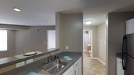 This single-floor residential unit offers a practical and well-organized layout designed for comfortable everyday living. The kitchen serves as a central feature, equipped with a double-basin stainless steel sink, dark countertops, and white cabinetry that provides ample storage. A breakfast bar or pass-through counter connects the kitchen to the adjacent living area, creating an open-concept flow that enhances the sense of space. The neutral gray wall tones carry consistently throughout the unit, lending a clean and cohesive aesthetic. A window in the living area allows natural light to filter in, brightening the communal spaces. The bathroom, visible from the main living area, features a shower with a curtain, a vanity with storage, and tile flooring, offering a functional and tidy design. The flooring in the main areas consists of durable tile, well-suited for high-traffic zones. Ceiling-mounted flush lighting fixtures are installed throughout, ensuring consistent illumination in each area. Overall, the unit presents a straightforward, move-in-ready layout with a neutral palette that accommodates a variety of interior styles.