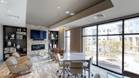This single-floor library is a well-appointed communal space designed to balance productivity and comfort. The open layout integrates a conference-style table with rolling chairs at its center, making it suitable for meetings, collaborative work, or focused study sessions. Adjacent to the table, a relaxed lounge area features mid-century modern accent chairs arranged around a glass coffee table, offering a more informal setting for reading or conversation.

A striking stone-clad accent wall anchors the lounge area, housing a sleek linear fireplace and a wall-mounted flat-screen television above it. Built-in shelving flanks the feature wall, providing organized display space for books and decorative objects. The flooring throughout combines wood-look planks with an area rug beneath the lounge seating, adding warmth and visual distinction between zones.

Floor-to-ceiling windows along one wall flood the interior with abundant natural light and frame views of the surrounding outdoor courtyard and landscaping. Recessed ceiling lighting and integrated speakers complement the modern, neutral color palette of taupe, gray, and warm beige tones, contributing to a polished and inviting ambiance.