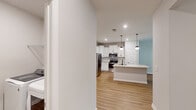 Located on Redbay Road in Simpsonville, South Carolina, this single-floor residence offers a thoughtfully designed layout with clean, contemporary finishes. The kitchen serves as a central highlight, featuring white shaker-style cabinetry extending to the upper walls, light granite countertops, a tiled backsplash, and a full suite of stainless steel appliances including a refrigerator, electric range, microwave, and dishwasher. A kitchen island with a granite surface and undermount sink provides additional prep and seating space, while two pendant lights add a polished touch. Light hardwood-style flooring runs throughout the main living and kitchen areas, complementing the neutral wall tones. The living area benefits from natural light through French doors topped with a transom window, offering views of surrounding greenery. Bedrooms feature plush carpeting and ceiling fans, with one bedroom providing direct access to a private bathroom fitted with a white vanity and granite countertop. A dedicated laundry room houses a full-size top-load washer and dryer alongside wire shelving for storage. The overall aesthetic is bright, clean, and move-in ready.