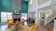 This clubhouse offers a thoughtfully designed, single-story community space that makes a strong impression from the moment you step through its welcoming entryway. The foyer is bright and inviting, featuring warm wood-look flooring, recessed lighting, and a decorative accent table that sets a polished tone for the rest of the space.

The main lounge area is the centerpiece of the clubhouse, defined by dramatically soaring double-height ceilings and a bold teal accent wall that draws the eye upward. A floor-to-ceiling tiled fireplace serves as the focal point, flanked by large windows that invite natural light and views of the surrounding community. Comfortable seating in warm amber and gold tones is arranged around rustic round tables, creating an ideal environment for relaxed socializing.

Adjacent to the lounge, a dedicated workspace area provides desks and chairs for residents seeking a productive setting. Abstract wall art adds a creative, contemporary flair throughout. The space transitions seamlessly into a more open, multi-purpose area with vaulted ceilings, abundant natural light from multiple windows, and direct access to the outdoors. Fitness equipment is also visible in this area, indicating a wellness component within the clubhouse. The consistent wood-look flooring and neutral wall palette unify the entire space with a clean, modern aesthetic.