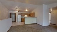 This single-floor space offers an open-concept design that seamlessly connects the living area and kitchen. The living area is finished with wall-to-wall carpeting, creating a warm and comfortable feel, while the kitchen features durable tile-style flooring. A peninsula counter separates the two areas, providing a natural division while maintaining the open flow between spaces.

The kitchen is equipped with medium-toned wood cabinetry offering generous upper and lower storage, a white refrigerator, and a range with an overhead ventilation hood. Recessed lighting illuminates the kitchen, complementing the flush-mount light fixture in the adjacent living area.

A notable architectural feature is the vaulted ceiling, which adds a sense of height and openness to the space. A ceiling fan with a light kit is centrally positioned in the living area, enhancing both air circulation and ambient lighting. Neutral white walls throughout provide a clean, versatile backdrop. A doorway visible in the background hints at additional rooms or storage, including what appears to be a closet with built-in shelving. The overall ambiance is functional, clean, and move-in ready.