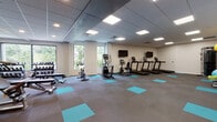 Located at 404 Algoma St in Madison, Wisconsin, this single-floor fitness center is designed to support a wide range of exercise needs in a clean, modern environment. The main workout area is generously sized and features a drop ceiling with recessed panel lighting that evenly illuminates the space. Distinctive teal accent squares are laid into the gray carpeted flooring throughout, lending a contemporary aesthetic to the facility.

The cardio zone is equipped with multiple treadmills, an elliptical trainer, and a stationary bike, all positioned near large windows that offer natural light and views of surrounding greenery. Wall-mounted flat-screen televisions accompany the cardio machines for an engaging workout experience. A dedicated free weights area includes a full dumbbell rack and weight benches, catering to strength training routines.

A functional training room provides additional space with a cable machine, medicine balls, stability balls, resistance bands, and rolling foam cylinders — ideal for dynamic and rehabilitative workouts. An adjacent mirrored studio area with full-length wall mirrors offers an open, reflective environment suited for stretching, yoga, or group fitness. A water fountain is conveniently accessible near the studio entrance, rounding out a comprehensive and well-appointed fitness facility.