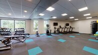 Located at 404 Algoma St in Madison, Wisconsin, this single-floor fitness center is designed to support a wide range of exercise needs in a clean, modern environment. The main workout area is generously sized and features a drop ceiling with recessed panel lighting that evenly illuminates the space. Distinctive teal accent squares are laid into the gray carpeted flooring throughout, lending a contemporary aesthetic to the facility.

The cardio zone is equipped with multiple treadmills, an elliptical trainer, and a stationary bike, all positioned near large windows that offer natural light and views of surrounding greenery. Wall-mounted flat-screen televisions accompany the cardio machines for an engaging workout experience. A dedicated free weights area includes a full dumbbell rack and weight benches, catering to strength training routines.

A functional training room provides additional space with a cable machine, medicine balls, stability balls, resistance bands, and rolling foam cylinders — ideal for dynamic and rehabilitative workouts. An adjacent mirrored studio area with full-length wall mirrors offers an open, reflective environment suited for stretching, yoga, or group fitness. A water fountain is conveniently accessible near the studio entrance, rounding out a comprehensive and well-appointed fitness facility.