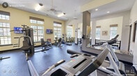 This fitness center in Raleigh, North Carolina offers a thoughtfully designed workout environment spread across a single, open floor. The space is divided into two functional zones, accommodating a wide range of exercise needs. Warm yellow-toned walls and durable rubber flooring set the tone throughout, while ceiling fans and recessed lighting ensure comfort during workouts.

Large multi-pane windows line the walls, flooding the interior with natural light and providing pleasant outdoor views of the surrounding community and greenery. The generous ceiling height enhances the sense of openness, making the space feel airy and inviting.

The main exercise area is well-stocked with cardio equipment, including multiple treadmills and elliptical machines, alongside strength training stations and cable machines. A wall of mirrors on one side visually expands the room and allows users to monitor their form. Medicine balls and stability equipment are also available for functional training.

A secondary zone, separated by a decorative metal railing, features additional specialty machines and open workout space. Wall-mounted televisions are positioned throughout for entertainment during cardio sessions. Safety features, including fire suppression equipment and clearly marked exits, are conveniently integrated into the layout. The overall design balances practicality with a welcoming atmosphere suited for daily fitness routines.
