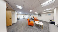 Located in Cambridge, MA, this well-appointed single-floor space offers a thoughtfully designed layout that blends comfort and functionality. The main open area is bright and welcoming, featuring a drop tile ceiling with recessed panel lighting that evenly illuminates the room throughout. Plush grey carpet tiles cover the floor, adding warmth and texture to the contemporary interior.

The lounge area is furnished with vibrant orange accent chairs and a grey sofa arranged around a sleek oval coffee table with gold-tone legs, creating an inviting social hub. Framed artwork adorns the neutral white walls, contributing to a polished, professional atmosphere. Large glass-paneled entry doors allow natural light to flow into the space, enhancing its open feel.

A dedicated workspace area features a tall standing workstation with adjustable stools, ideal for collaborative use. Adjacent to this, a kitchenette with a refrigerator, countertop workspace, and dining table with chairs provides practical amenities for occupants. A hallway connects to the full bathroom, which is finished with neutral floor tiles, a wall-mounted vanity, and commercial-grade fixtures. The overall design balances modern aesthetics with practical, multi-use functionality, making it well-suited as a communal or leasing office environment.