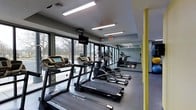 This fitness center is designed to deliver a functional and motivating workout experience within a clean, contemporary setting. The space is anchored by a generous row of cardio equipment, including treadmills and elliptical machines, arranged to maximize floor space and ease of movement between stations. Floor-to-ceiling windows line one wall, flooding the interior with natural light and offering open views of the surrounding outdoor landscape, creating a bright and refreshing atmosphere during workouts. Large mirrors span an adjacent wall, visually expanding the space and allowing users to monitor their form. Overhead, recessed linear fluorescent lighting provides consistent, even illumination throughout. The flooring is finished in a durable grey tone suited for high-traffic fitness use. Accent yellow structural columns add a bold visual contrast to the otherwise neutral palette of grey and black. A wall-mounted television is positioned for convenient viewing from the cardio machines. The overall ambiance is modern and energetic, with an emphasis on openness, functionality, and comfort — making it a well-appointed facility for a variety of fitness routines.