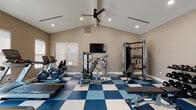This dedicated fitness center is thoughtfully designed to support a comprehensive workout experience. The space is anchored by striking blue-and-white checkered flooring that adds a bold visual element throughout. Vaulted ceilings rise overhead, creating an open, airy atmosphere enhanced by recessed lighting and a ceiling fan that keeps the environment comfortable during exercise.

The cardio zone lines one wall and features multiple Matrix treadmills and upright stationary bikes, positioned in front of large windows fitted with roller shades that allow natural light to filter in. A wall-mounted flat-screen television is centrally positioned, providing entertainment during workouts.

The strength training area is well-appointed with a multi-function cable machine, a dedicated weight bench, and a power rack equipped with kettlebells and additional attachments. A three-tier dumbbell rack holds a full range of free weights, offering versatility for various training routines. Additional machines support targeted muscle group workouts, ensuring users have access to both functional and machine-based training options.

The neutral warm-toned walls provide a clean backdrop that keeps the focus on the equipment. Natural light enters through multiple windows, contributing to an energizing atmosphere. The layout is organized and spacious, allowing comfortable movement between stations throughout the entire fitness floor.