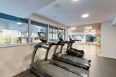 Located in Vancouver's West End neighbourhood, this residential fitness centre offers a thoughtfully designed, two-zone layout across a single floor. The space is divided into a fully equipped gym area and a separate multi-purpose studio, connected by a glass-partitioned wall and door that allows natural light to flow between zones while maintaining visual separation.

The cardio and strength training area features rubberized flooring and houses a lineup of TRUE-brand treadmills and ellipticals positioned along a bank of windows, providing natural light and views of the surrounding neighbourhood. Strength training equipment, including cable machines, weight benches, and free weights, occupies an adjacent section. Recessed ceiling lights ensure bright, even illumination throughout.

The multi-purpose studio is a standout space, finished with warm hardwood flooring and a dark ceiling that creates a refined, focused atmosphere. Large windows wrap the room, flooding it with natural light. Full-length mirrors along one wall visually expand the space, making it ideal for yoga, stretching, or group classes. Exercise balls, resistance tools, dumbbells, and a mounted flat-screen TV complete the studio's amenities. Textured wood-panelled accent walls add warmth and a contemporary design touch throughout both zones.