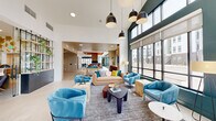 Located at 6609 E Warren Dr. in Denver, CO, this thoughtfully designed single-floor amenity space offers an expansive and welcoming environment for residents. The open-concept living and lounge area immediately impresses with soaring double-height ceilings, floor-to-ceiling windows, and an abundance of natural light that floods the interior. Bold teal accent walls contrast beautifully with warm wood tones and light hardwood flooring throughout, creating a contemporary yet inviting atmosphere. The lounge is furnished with vibrant blue upholstered seating, layered area rugs, and statement pendant lighting, while a large built-in media wall with a multi-screen display serves as a central focal point. A pool table adds a recreational element to the space. The kitchen features sleek light wood cabinetry with brass hardware, granite countertops, and a striking floor-to-ceiling teal tile backsplash. Glass doors open directly to an outdoor area, seamlessly connecting the interior to the exterior. The dining area offers cozy booth-style seating nestled within a glass-wrapped nook, bathed in natural light. A private dining or meeting room with rich teal walls and curated artwork provides a more intimate setting. Three connecting hallways ensure smooth circulation throughout this generously proportioned space.