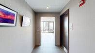 This elevated amenity space is designed to offer both refined indoor comfort and striking outdoor living. Entry is made through a clean, modern corridor finished with large-format tile flooring, neutral taupe walls, recessed lighting, and rich dark wood accent doors — setting a polished tone from the outset.

The interior lounge area is bright and contemporary, characterized by floor-to-ceiling glass walls that flood the space with natural light and frame sweeping views of the surrounding cityscape. The lounge is appointed with a comfortable seating arrangement, a wall-mounted flat-screen television, and large-scale artwork that adds a coastal aesthetic. Recessed and track lighting complement the abundant daylight throughout.

Adjacent to the lounge is a well-equipped kitchenette featuring wood cabinetry, a white tile backsplash, stainless steel appliances, and a stylish dining table with pendant lighting overhead — ideal for gatherings and events.

The crowning feature of this space is the expansive rooftop terrace, accessible directly from the interior. The terrace is furnished with teak and upholstered outdoor seating arrangements and is enclosed by a sleek glass railing system, ensuring unobstructed panoramic views of the city skyline, lush green treetops, and a glistening body of water in the distance. The open-air setting is bathed in sunlight, creating an exceptional environment for relaxation and socializing.