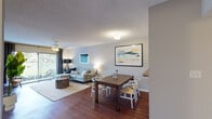 This single-floor residence offers a thoughtfully designed open-concept layout that seamlessly connects the living, dining, and kitchen areas. Rich hardwood flooring flows throughout the main living spaces, creating a warm and inviting ambiance. The living room is generously proportioned, anchored by a tufted sectional sofa, ceiling fan, and a large sliding glass door that opens to an outdoor balcony, flooding the space with natural light and offering views of lush greenery. Adjacent to the living area, the dining space comfortably accommodates a full dining table and chairs, with easy access to the kitchen. The kitchen features dark cabinetry, granite-style countertops, and black appliances, with tile flooring extending through the functional galley-style corridor. The bedroom is carpeted for comfort, equipped with a ceiling fan, a large window overlooking mature trees, and ample space for a full bedroom suite. The bathroom includes a dark wood vanity, a wide mirror with overhead lighting, and a shower-tub combination. Throughout the residence, a textured ceiling, neutral wall tones, and consistent design choices contribute to a cohesive and comfortable living environment.