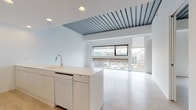 Located on Euclid Avenue in Cleveland's University Circle neighborhood, this single-floor unit offers a clean, modern aesthetic defined by an all-white palette and thoughtfully selected finishes. The open-concept living and kitchen area serves as the heart of the space, anchored by a large white island with an integrated sink and ample counter seating. The kitchen is fully equipped with high-gloss white cabinetry extending to the ceiling, a stainless steel refrigerator, built-in oven, and microwave. Recessed lighting illuminates the space throughout. A defining architectural feature is the exposed corrugated metal ceiling detail in the living area, which adds industrial character and visual contrast to the otherwise minimalist interior. Floor-to-ceiling windows flood the living space with natural light and frame sweeping views of the Cleveland cityscape. Light wood-tone flooring runs through the bedroom areas, while polished concrete floors anchor the main living zone. The bathroom is finished with white square mosaic tile on both the floor and walls, complemented by a soaking tub with shower combination and a glass partition. The bedroom offers generous wall space and tall ceilings, with direct access to the bathroom. This unit combines urban design sensibility with functional living spaces in a prime Cleveland location.