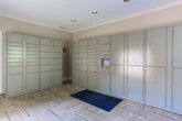 Located at 80 Newnan Lakes Blvd in Newnan, Georgia, this single-floor package locker room is a dedicated parcel management facility designed for convenience and security. The room is lined with rows of floor-to-ceiling secure package lockers finished in a neutral gray tone, equipped with a digital touchscreen interface for easy access and retrieval. Crown molding details along the ceiling add a polished architectural touch to the otherwise functional space. The flooring consists of patterned carpet tiles in a warm neutral palette, accented by a bold blue entryway mat at the door threshold. Natural light floods the interior through large windows fitted with horizontal blinds and complemented by decorative blue patterned curtain panels, offering pleasant views of the surrounding greenery outside. The exterior-facing glass-panel door provides an additional point of entry and exit, clearly marked with illuminated exit signage for safety compliance. Recessed ceiling lighting ensures the room remains well-lit at all times. The warm beige walls and crown detailing create a welcoming ambiance, balancing the room's practical purpose with a refined aesthetic consistent with a well-maintained residential community.