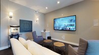 Located at 5793 Hoover Falls Dr in Westerville, Ohio, this single-floor home offers a thoughtfully designed living experience with a standout entertainment space at its heart. The media room is a highlight of the home, centered around a large wall-mounted flat-screen television and outfitted with comfortable, coordinated seating including a plush sofa and accent chairs in soft gray tones. Hexagonal wooden ottomans add a modern, geometric touch to the room's aesthetic. Recessed lighting and wall sconces provide layered illumination, creating a warm and immersive atmosphere ideal for movie nights or casual gatherings. A film reel motif on the wall reinforces the cinematic theme of the space, while a compact built-in cabinet offers convenient storage. Neutral wall tones and dark carpeting contribute to the cozy, theater-like ambiance. The room's design reflects a careful balance of function and style, combining entertainment-focused features with tasteful décor elements. The overall layout and finishes of this home suggest a modern, well-appointed residence designed for comfortable everyday living and quality leisure time.