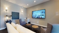 Located at 5793 Hoover Falls Dr in Westerville, Ohio, this single-floor home offers a thoughtfully designed living experience with a standout entertainment space at its heart. The media room is a highlight of the home, centered around a large wall-mounted flat-screen television and outfitted with comfortable, coordinated seating including a plush sofa and accent chairs in soft gray tones. Hexagonal wooden ottomans add a modern, geometric touch to the room's aesthetic. Recessed lighting and wall sconces provide layered illumination, creating a warm and immersive atmosphere ideal for movie nights or casual gatherings. A film reel motif on the wall reinforces the cinematic theme of the space, while a compact built-in cabinet offers convenient storage. Neutral wall tones and dark carpeting contribute to the cozy, theater-like ambiance. The room's design reflects a careful balance of function and style, combining entertainment-focused features with tasteful décor elements. The overall layout and finishes of this home suggest a modern, well-appointed residence designed for comfortable everyday living and quality leisure time.