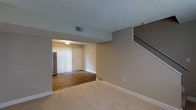 This two-story residence offers a straightforward and functional layout designed for comfortable everyday living. The main level features a spacious carpeted living area with neutral beige walls and a textured ceiling, creating a clean and understated atmosphere. A central opening connects the living space to the entryway, which is finished with warm hardwood flooring and provides direct access to the front door. Adjacent to the entry, a refrigerator is partially visible, indicating a kitchen or kitchenette in close proximity. HVAC vents are positioned throughout, ensuring climate control across the space. A staircase with white-painted railings rises along one wall, leading to the second floor and adding a sense of verticality to the interior. The consistent use of neutral tones — from the walls to the flooring — gives the space a cohesive and versatile aesthetic, making it easy to personalize. Natural light filters through vertical blinds near the entryway, contributing to a bright and welcoming feel. Overall, this two-level space combines practical design with a calm, neutral palette suited for a variety of living arrangements.